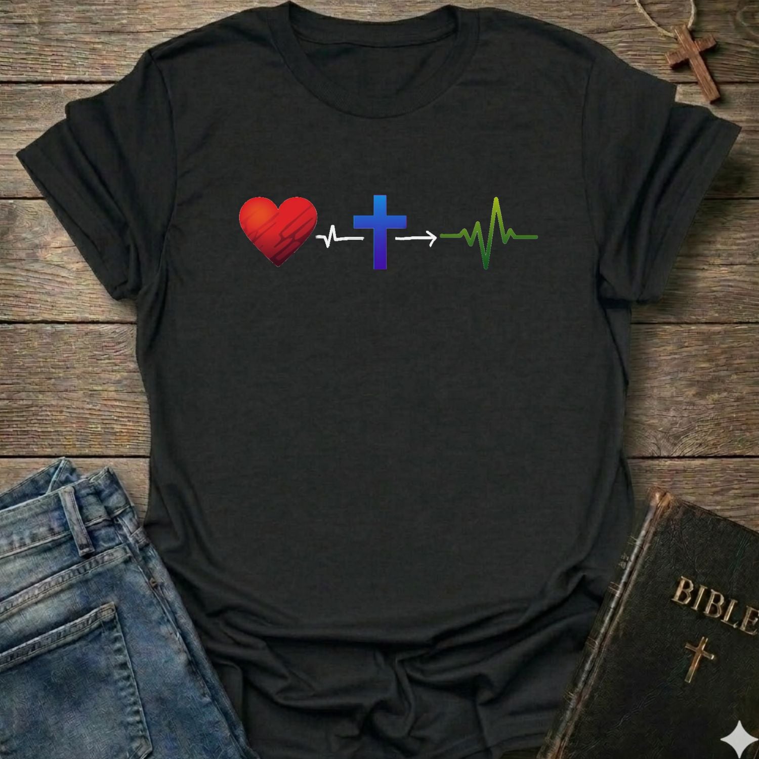 A black short-sleeve t-shirt laid flat on a rustic wooden surface, featuring a graphic of a red heart, a blue cross, and a green heartbeat line connected by arrows. Blue jeans and a Bible are partially visible as styling props.