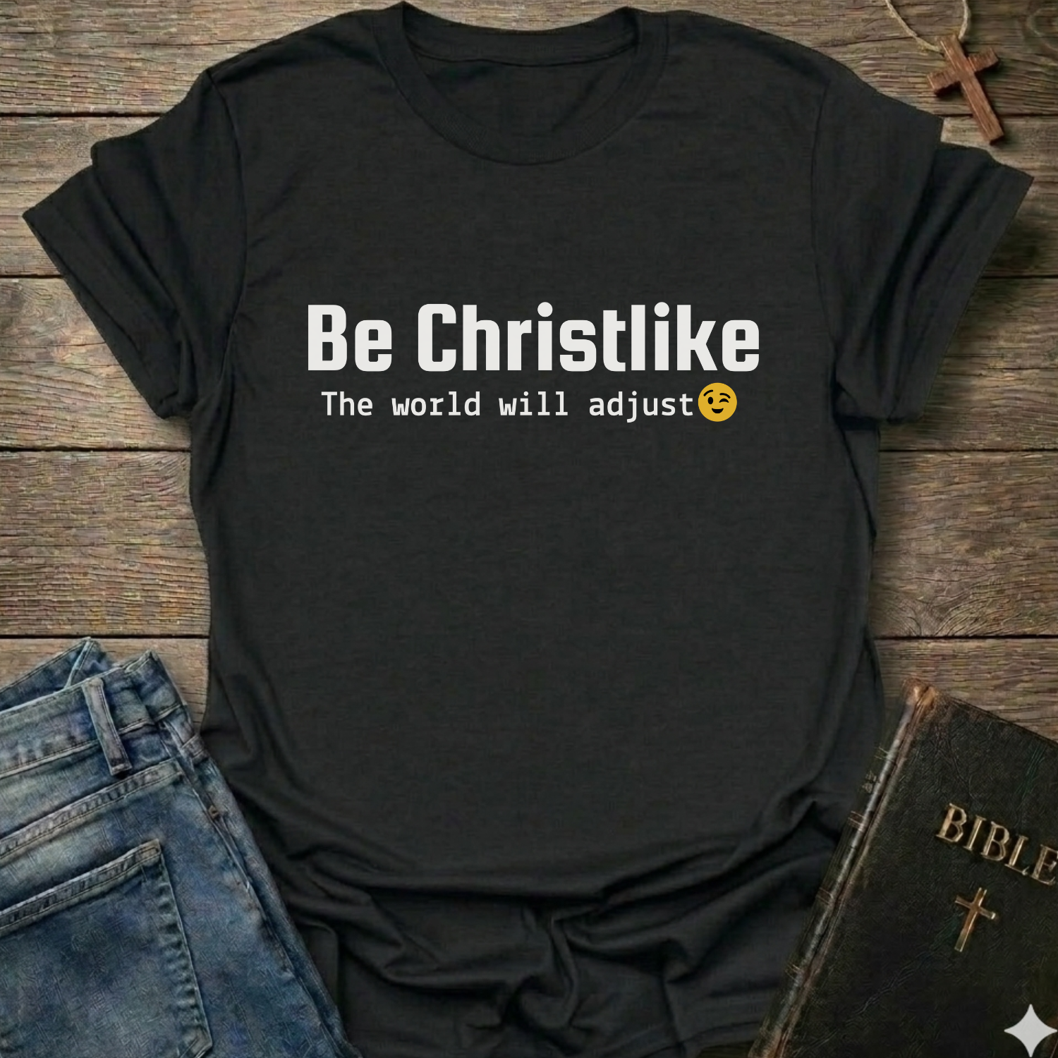Black t-shirt with 'Be Christlike The world will adjust' text on a wooden surface with jeans and a Bible.