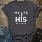Gray t-shirt with 'My Life Is His'  Christian T-Shirt Galatians 2:20 bold faith apparel Faith Forward text on a wooden surface with jeans and a Bible.