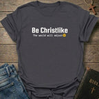 A dark heather gray t-shirt with the text 'Be Christlike The world will adjust' and a small yellow smiley face, styled in a flat-lay with blue jeans, a Bible, and a wooden cross on a rustic wood background.