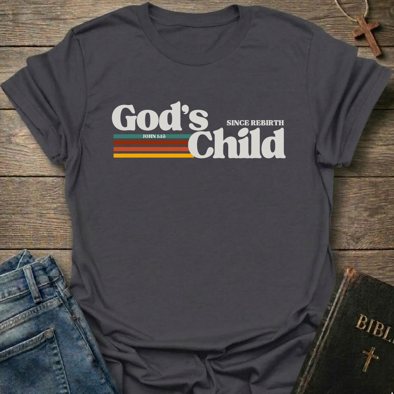 Gray t-shirt with God's Child Since Rebirth Christian T-Shirt John 1:12 retro faith apparel conversation starter text on a wooden surface with jeans and a Bible.