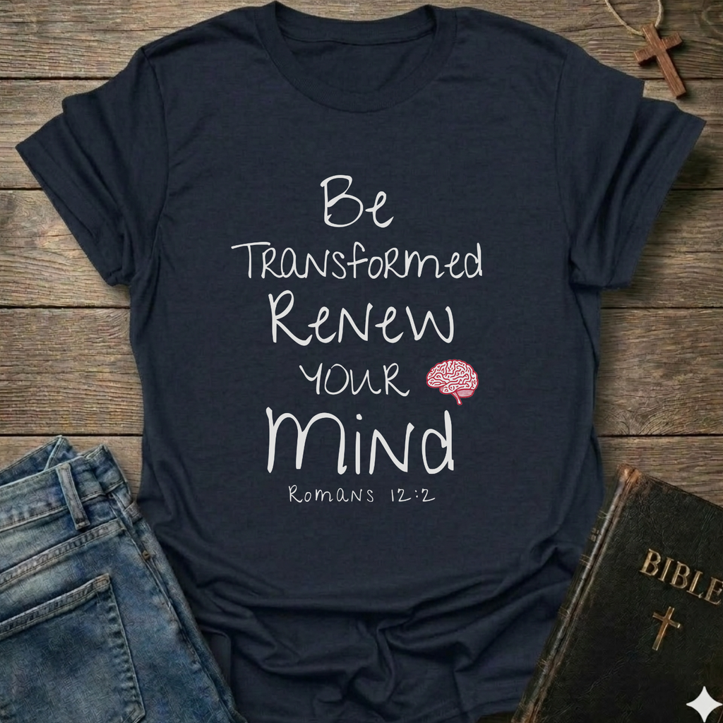 A navy blue graphic t-shirt laid flat on a rustic wooden background, styled with blue jeans, a Bible, and a wooden cross. The t-shirt features white text reading 'Be Transformed Renew Your Mind Romans 12:2' with a small red brain graphic.