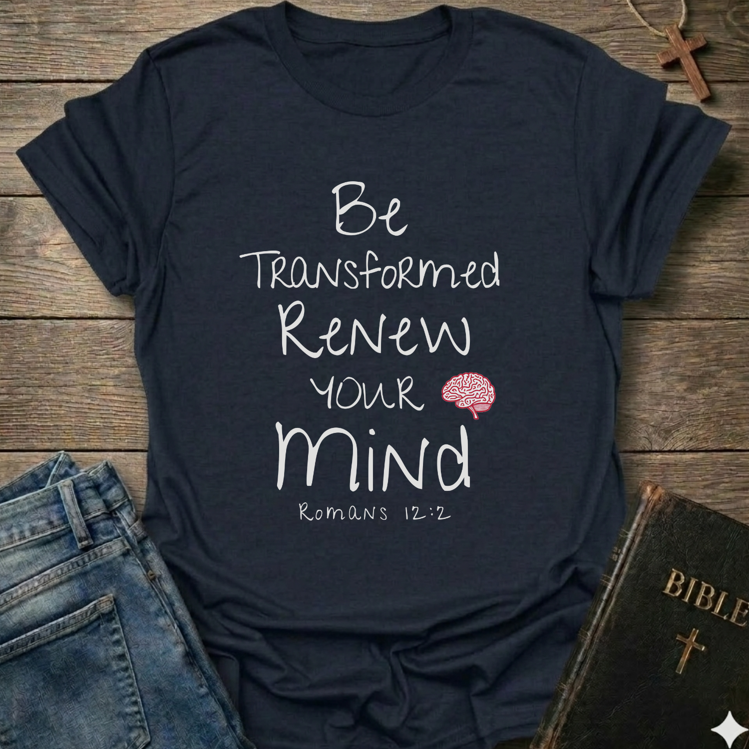 A navy blue graphic t-shirt laid flat on a rustic wooden background, styled with blue jeans, a Bible, and a wooden cross. The t-shirt features white text reading 'Be Transformed Renew Your Mind Romans 12:2' with a small red brain graphic.