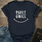 A navy blue graphic t-shirt with a white smiley face design and the text 'SMILE JESUS CARES' and 'SMILL' printed on the front. The shirt is styled in a flat-lay arrangement on a rustic wooden background, accompanied by blue jeans, a brown cross necklace, and a Bible.