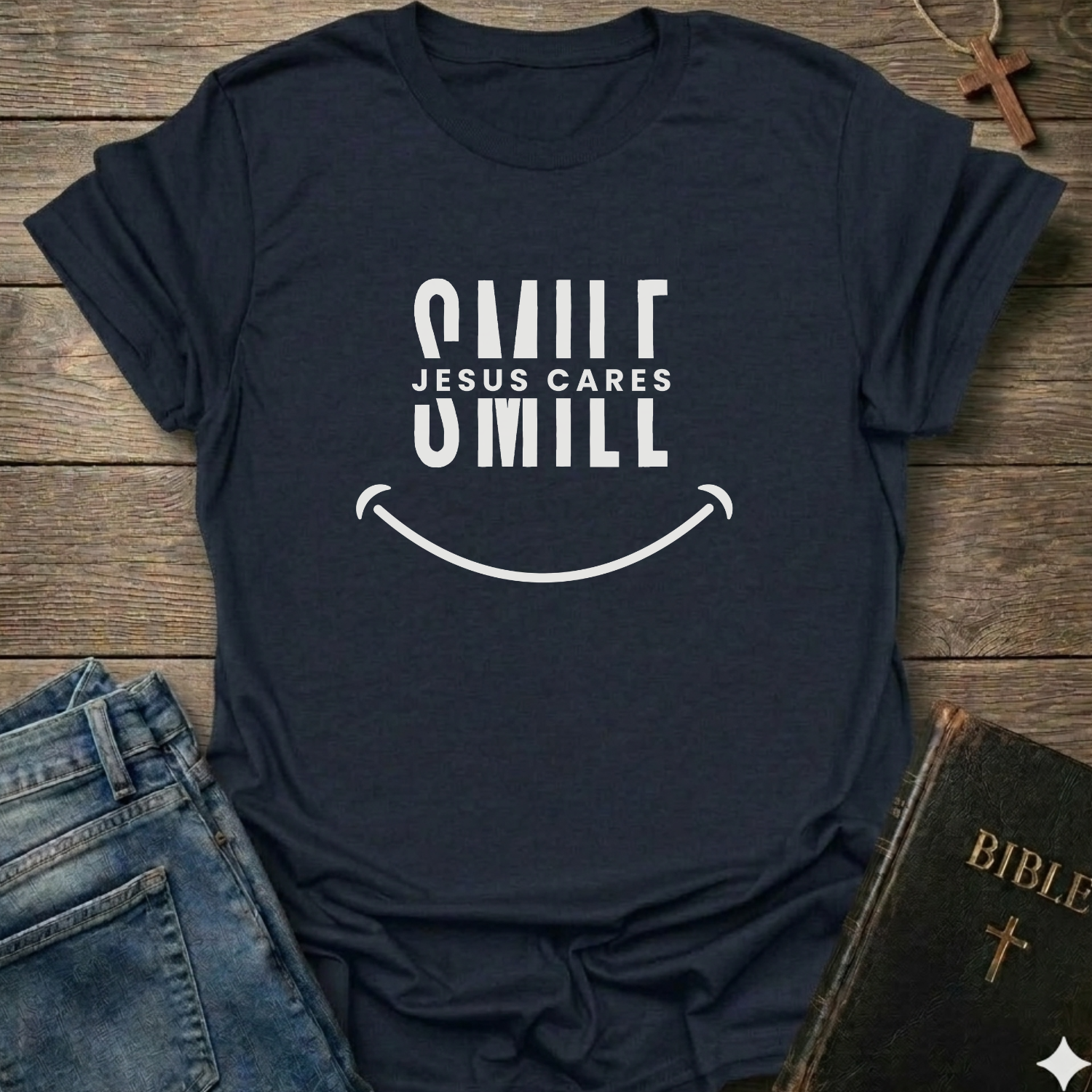 A navy blue graphic t-shirt with a white smiley face design and the text 'SMILE JESUS CARES' and 'SMILL' printed on the front. The shirt is styled in a flat-lay arrangement on a rustic wooden background, accompanied by blue jeans, a brown cross necklace, and a Bible.