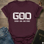 A maroon short-sleeve t-shirt with a white graphic print reading 'GOD IS FATHER • SON • HOLY SPIRIT', styled in a flat-lay on a rustic wooden background with blue jeans and a Bible nearby.
