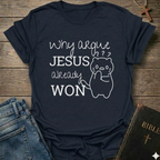 A navy blue graphic t-shirt laid flat on a rustic wooden background, styled with blue jeans on the left and a Bible on the right. The t-shirt features a white graphic print that reads 'Why argue JESUS already WON' alongside a simple line drawing of a cat with confused facial expressions.