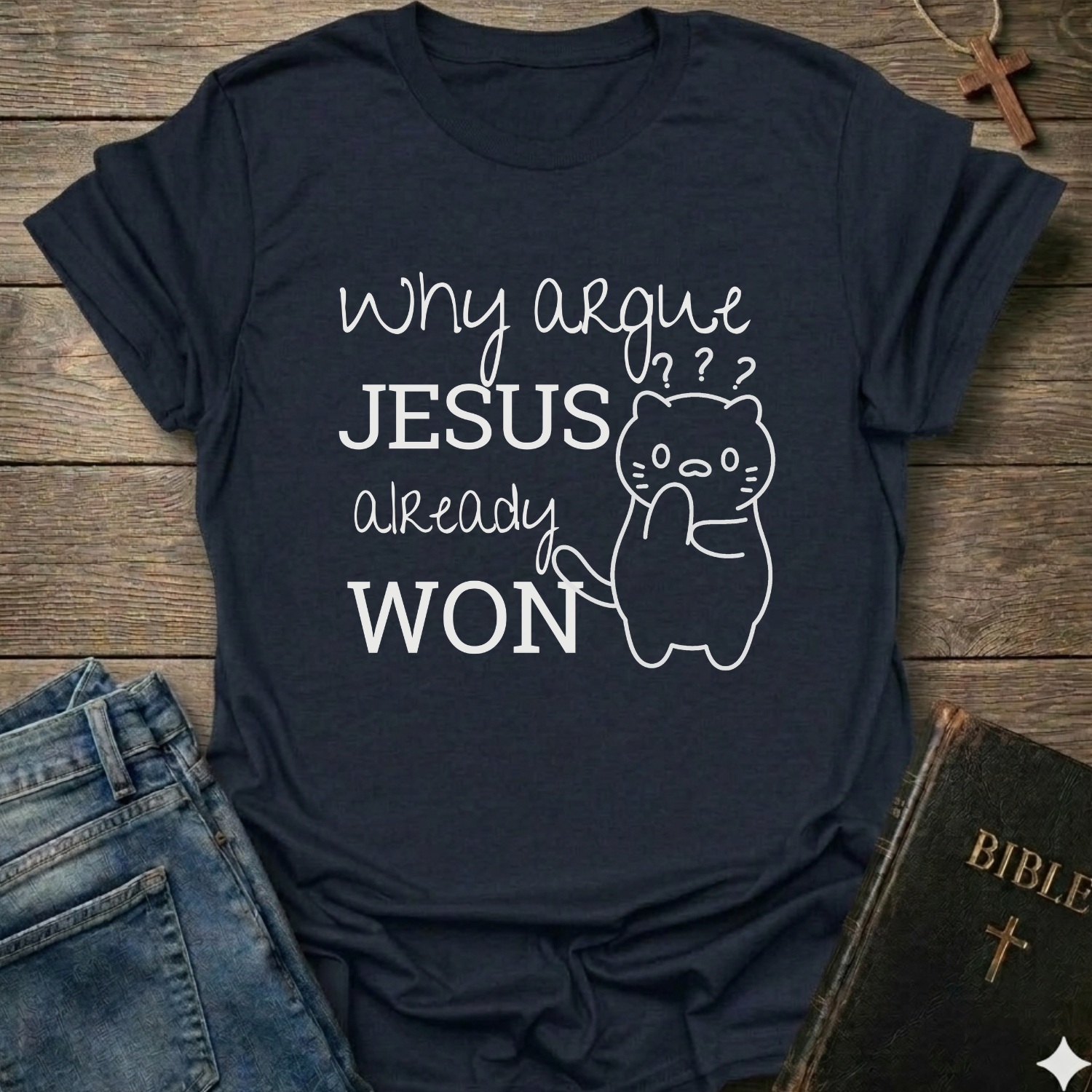 A navy blue graphic t-shirt laid flat on a rustic wooden background, styled with blue jeans on the left and a Bible on the right. The t-shirt features a white graphic print that reads 'Why argue JESUS already WON' alongside a simple line drawing of a cat with confused facial expressions.