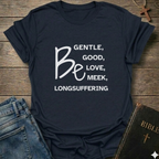 A navy blue graphic t-shirt with white text reading 'BE GENTLE, GOOD, LOVE, MEEK, LONGSUFFERING' styled in a flat-lay on a rustic wooden background with jeans and a Bible nearby.