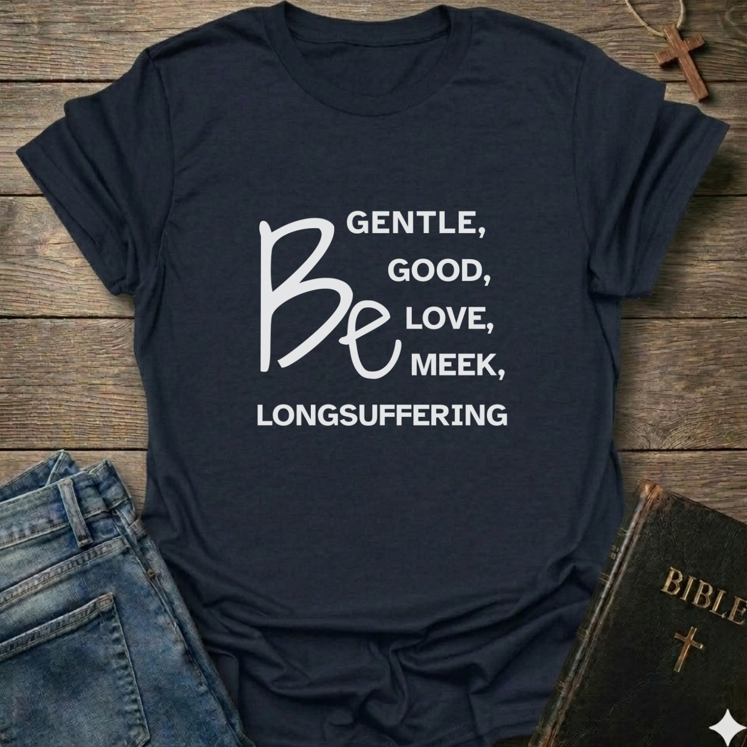A navy blue graphic t-shirt with white text reading 'BE GENTLE, GOOD, LOVE, MEEK, LONGSUFFERING' styled in a flat-lay on a rustic wooden background with jeans and a Bible nearby.