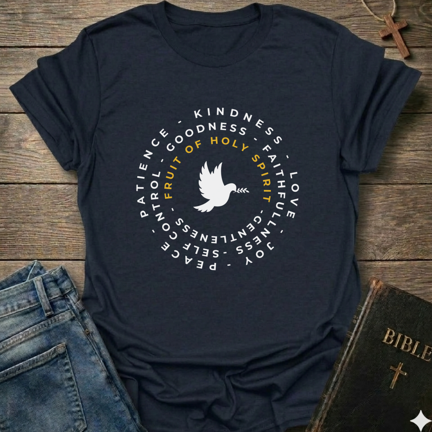A navy blue graphic t-shirt laid flat on a rustic wooden surface, styled with blue jeans, a Bible, and a wooden cross. The t-shirt features a white circular graphic with a dove in the center and the words 'Fruit of Holy Spirit' in yellow, surrounded by white words including 'PATIENCE', 'KINDNESS', 'GOODNESS', 'FAITH', 'LOVE', 'JOY', 'PEACE', and 'GENTLENESS'.