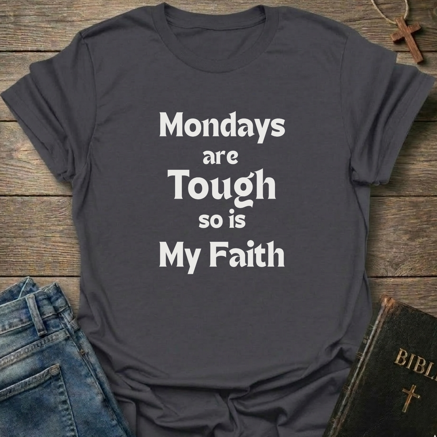 A heather dark gray graphic t-shirt with the text 'Mondays are Tough so is My Faith' printed in white. The t-shirt is styled in a flat-lay arrangement on a rustic wooden background, accompanied by blue jeans, a brown wooden cross, and a vintage-style Bible.