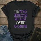 Black t-shirt with motivational text 'The yoke destroyed because of the anointing' Isaiah 10:27 faith apparel conversation starter on a wooden surface with jeans and a Bible.