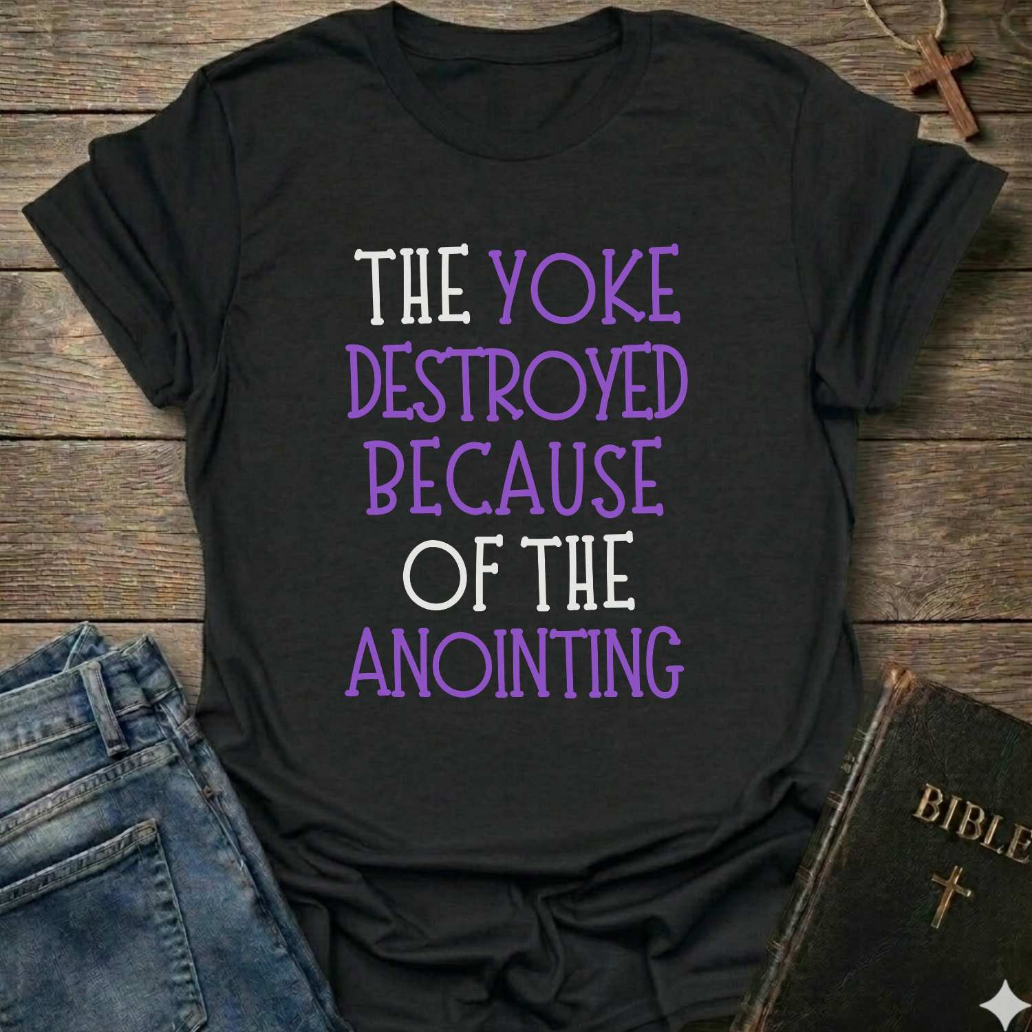 Black t-shirt with motivational text 'The yoke destroyed because of the anointing' Isaiah 10:27 faith apparel conversation starter on a wooden surface with jeans and a Bible.