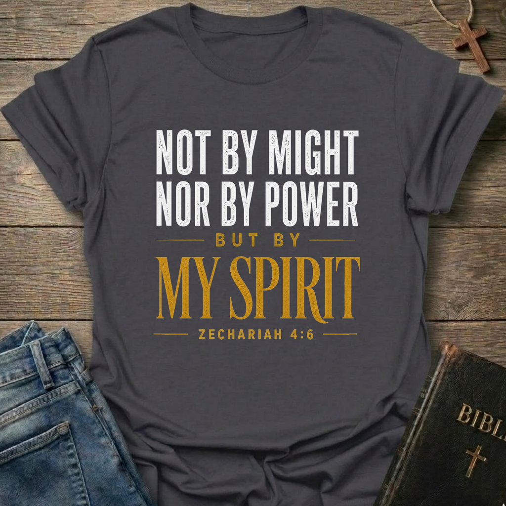 A dark charcoal gray graphic t-shirt laid flat on a rustic wooden background, styled with blue jeans, a leather Bible, and a wooden cross. The t-shirt features the text 'NOT BY MIGHT NOR BY POWER BUT BY MY SPIRIT ZECHARIAH 4:6' in white and gold lettering.
