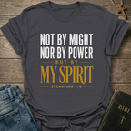 A dark charcoal gray graphic t-shirt laid flat on a rustic wooden background, styled with blue jeans, a leather Bible, and a wooden cross. The t-shirt features the text 'NOT BY MIGHT NOR BY POWER BUT BY MY SPIRIT ZECHARIAH 4:6' in white and gold lettering.