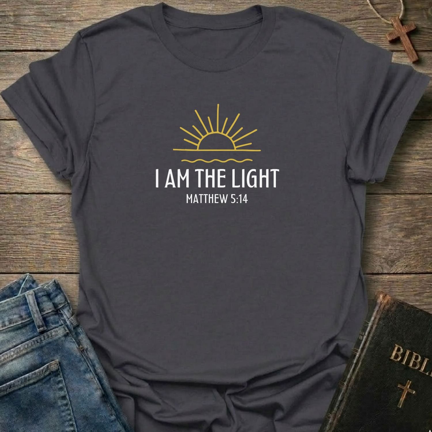 A dark heather gray t-shirt with a yellow and white graphic print reading 'I AM THE LIGHT MATTHEW 5:14' under a rising sun design. The shirt is styled in a flat-lay on a rustic wooden background, accompanied by blue jeans, a black Bible, and a wooden cross.
