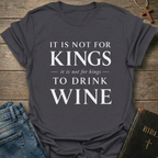 A dark heather gray graphic t-shirt with white text reading 'IT IS NOT FOR KINGS it is not for kings TO DRINK WINE', styled in a flat-lay on a rustic wooden background with blue jeans and a Bible nearby.