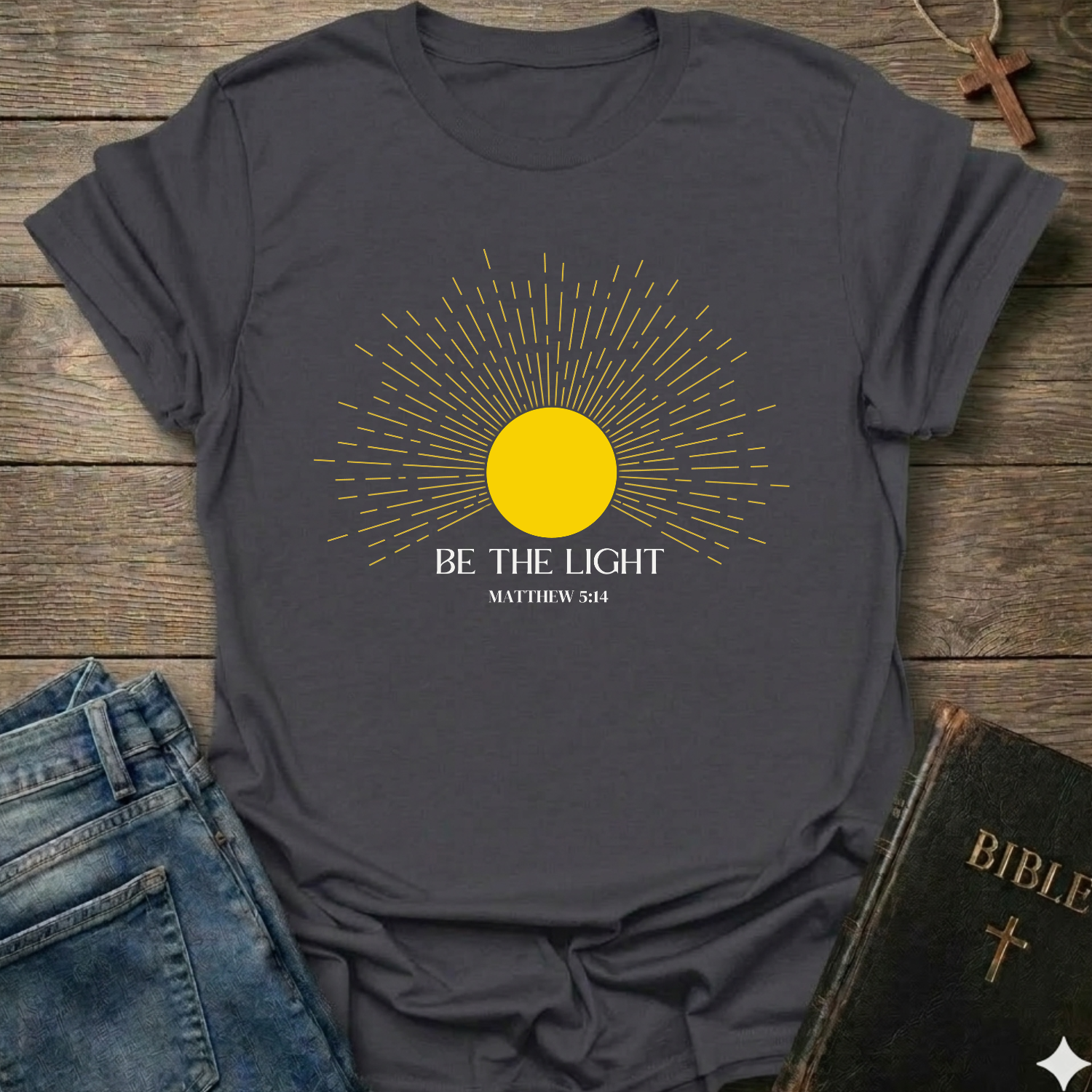 Gray t-shirt with yellow sun graphic and 'Be the Light' text on a wooden surface with jeans and a Bible. Be the Light Christian T-Shirt Matthew 5:14 bold faith apparel Faith Forward