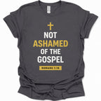 A dark heather gray short-sleeve t-shirt with a graphic print on the front. The print features a gold Christian cross, the text 'NOT ASHAMED OF THE GOSPEL' in white and gold, and a gold box containing 'ROMANS 1:16' at the bottom.