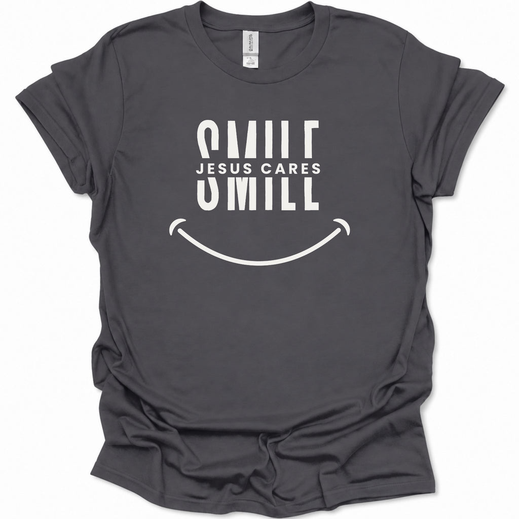 A dark heather gray short-sleeve t-shirt with a white graphic print that reads 'SMILE JESUS CARES SMILL' above a smiley face, shown on a plain white background.