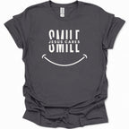 A dark heather gray short-sleeve t-shirt with a white graphic print that reads 'SMILE JESUS CARES SMILL' above a smiley face, shown on a plain white background.