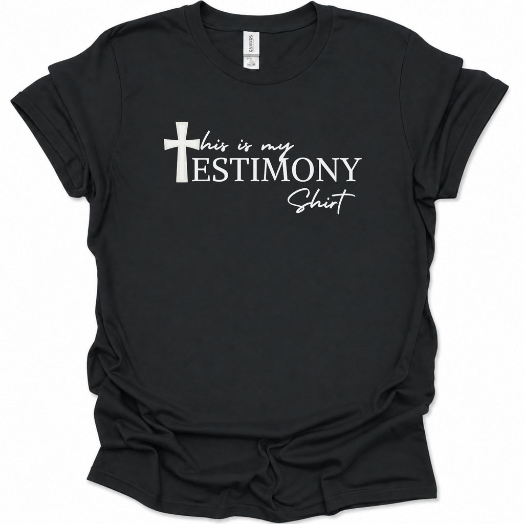 A black short-sleeve t-shirt with a white graphic print on the front that reads 'this is my TESTIMONY Shirt' alongside a Christian cross, displayed against a plain white background.