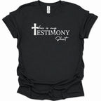 A black short-sleeve t-shirt with a white graphic print on the front that reads 'this is my TESTIMONY Shirt' alongside a Christian cross, displayed against a plain white background.