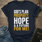 A navy blue graphic t-shirt laid flat on a rustic wooden background, styled with blue jeans, a Bible, and a wooden cross. The t-shirt features the text 'GOD'S PLAN: PROSPERITY, HOPE & A FUTURE FOR ME!' in bold white and yellow lettering.