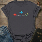 Gray t-shirt with a colorful heart, cross, and heartbeat line design on a wooden surface with jeans and a Bible.