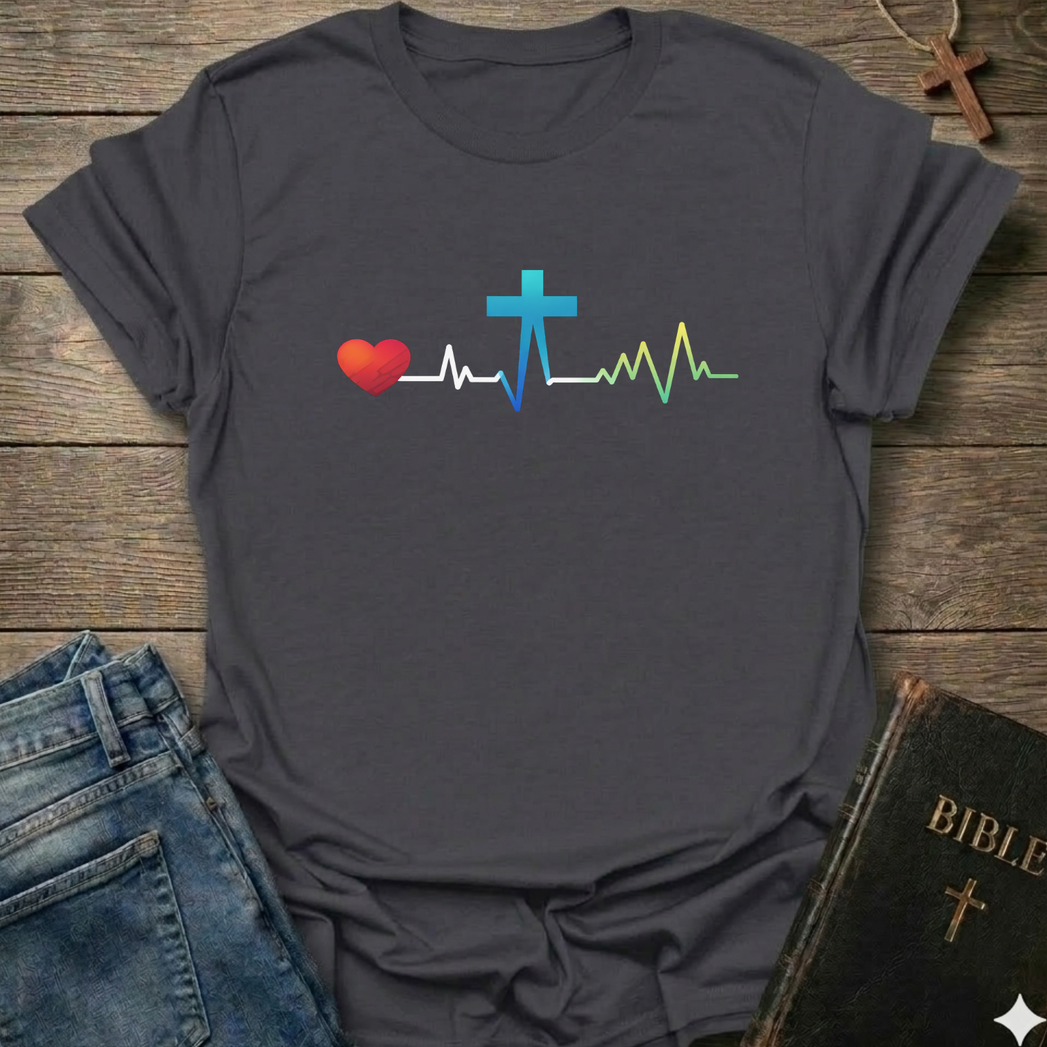 Gray t-shirt with a colorful heart, cross, and heartbeat line design on a wooden surface with jeans and a Bible.