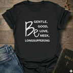 A black graphic t-shirt with white text reading 'BE GENTLE, GOOD, LOVE, MEEK, LONGSUFFERING' styled in a flat-lay on a rustic wooden background with blue jeans and a Bible nearby.
