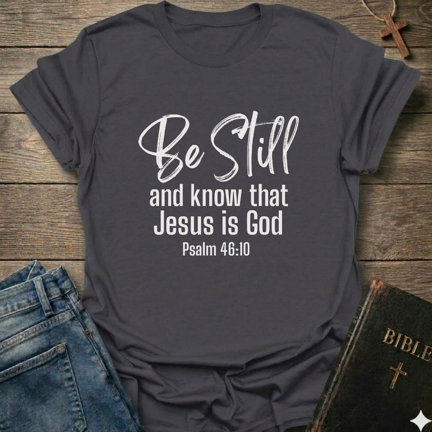 Gray t-shirt with 'Be Still and know that Jesus is God' text Psalm 46:10 script faith apparel Faith Forward
on a wooden surface with jeans and a Bible.
