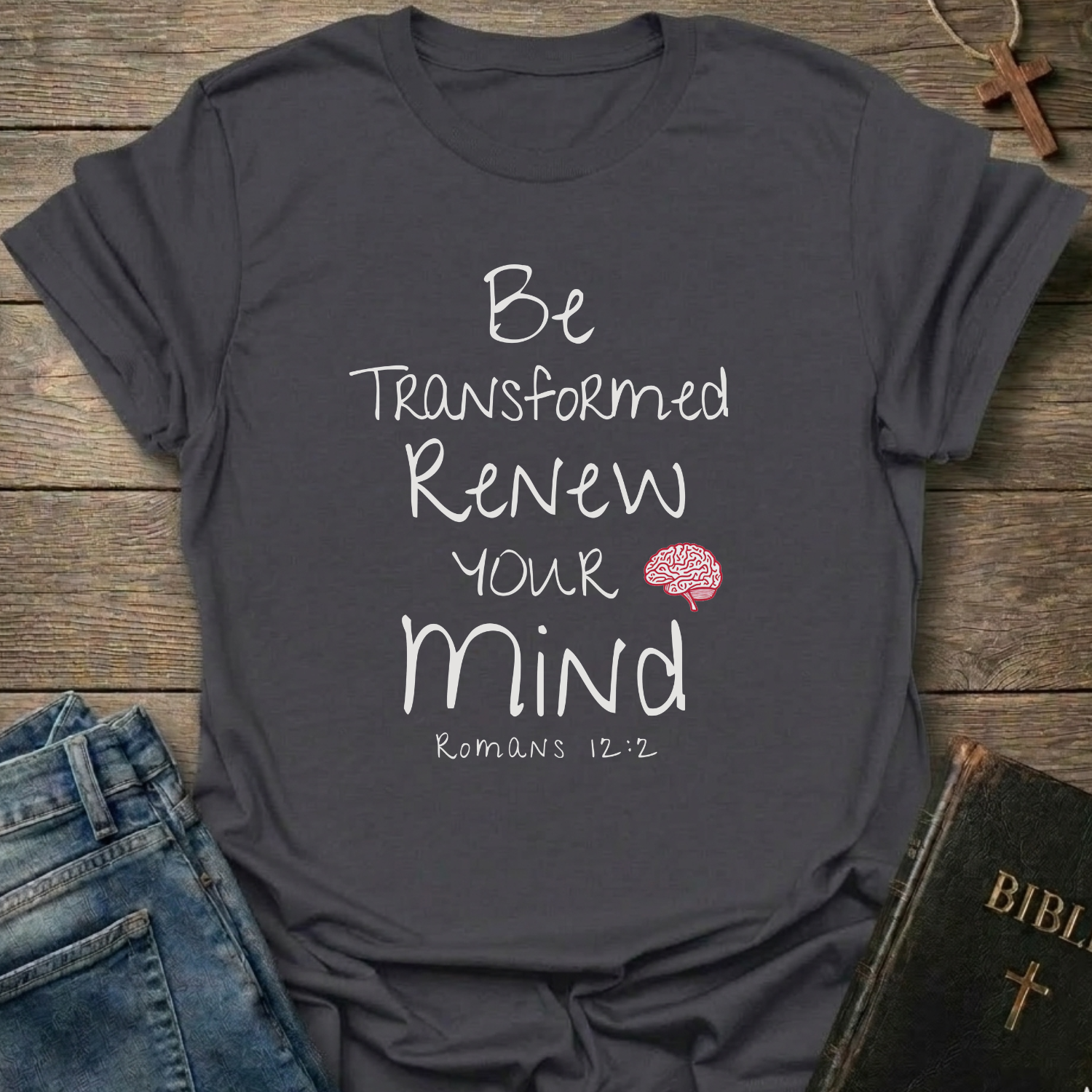 A dark charcoal gray graphic t-shirt laid flat on a rustic wooden background, styled with blue jeans, a Bible, and a wooden cross. The t-shirt features the text 'Be Transformed Renew Your Mind Romans 12:2' in white lettering with a small red brain illustration.