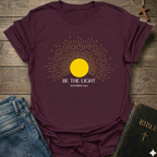 Maroon t-shirt with yellow sun graphic and 'Be the Light' text on a wooden surface with jeans and a Bible. Be the Light Christian T-Shirt Matthew 5:14 bold faith apparel Faith Forward