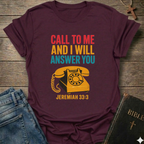 Maroon t-shirt with vintage phone graphic and text Call to Me and I Will Answer You Jeremiah 33:3 retro phone faith Christian apparel conversation starter on a wooden surface with jeans and a Bible.