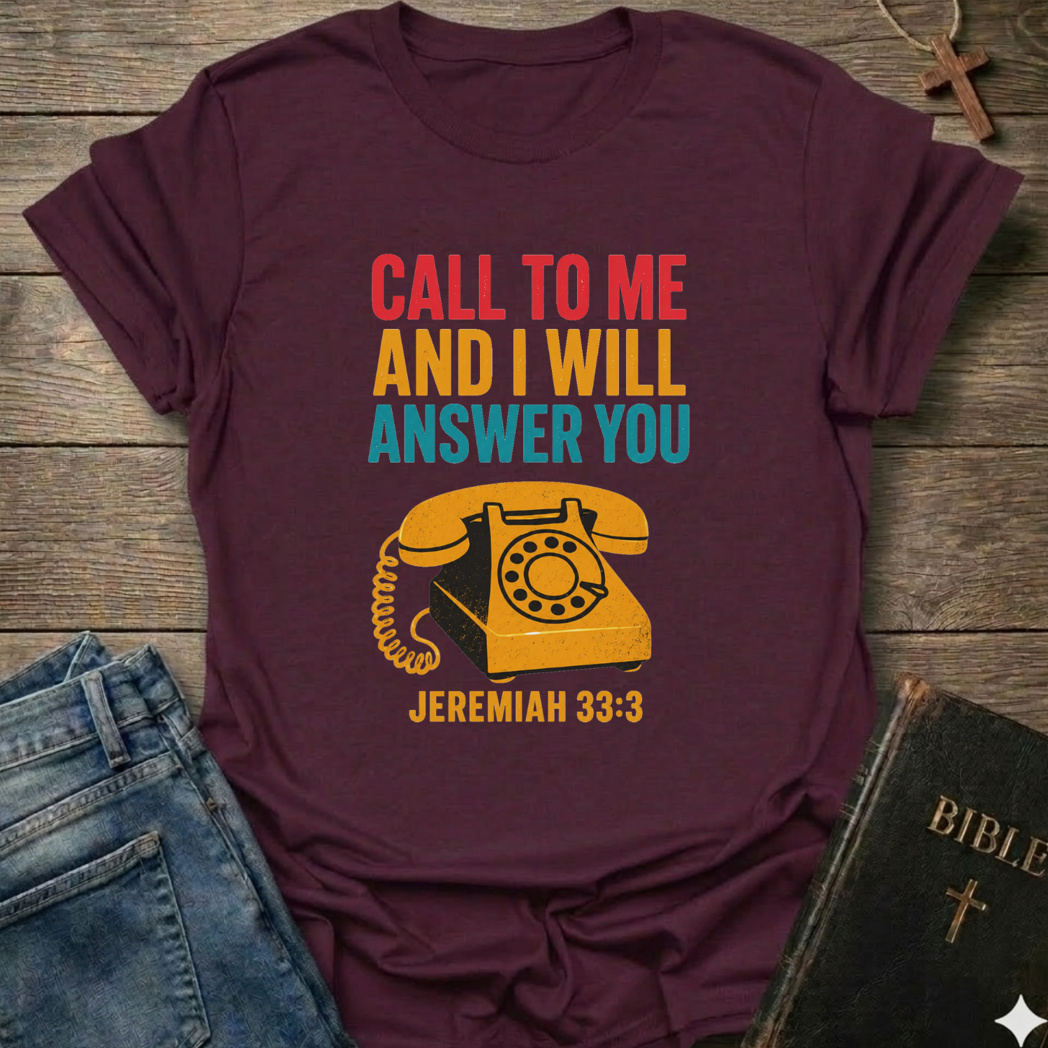Maroon t-shirt with vintage phone graphic and text Call to Me and I Will Answer You Jeremiah 33:3 retro phone faith Christian apparel conversation starter on a wooden surface with jeans and a Bible.