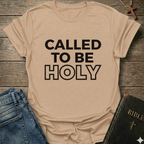 Natural t-shirt with 'Called to be Holy' Christian T-Shirt bold faith apparel 1 Peter 1:15 conversation starter text on a wooden surface with jeans and a Bible.