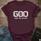 Maroon T-shirt with text "God is Father Son Holy Spirit Trinity" Christian T-Shirt bold typography faith apparel conversation starter. It is on a wooden background with a jeans and a bible.