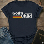 Navy blue t-shirt with God's Child Since Rebirth Christian T-Shirt John 1:12 retro faith apparel conversation starter text on a wooden surface with jeans and a Bible.
