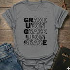 Gray t-shirt with 'GRACE UPON GRACE' text on a wooden surface with jeans and a Bible. Grace Upon Grace Christian T-Shirt John 1:16 bold faith apparel 