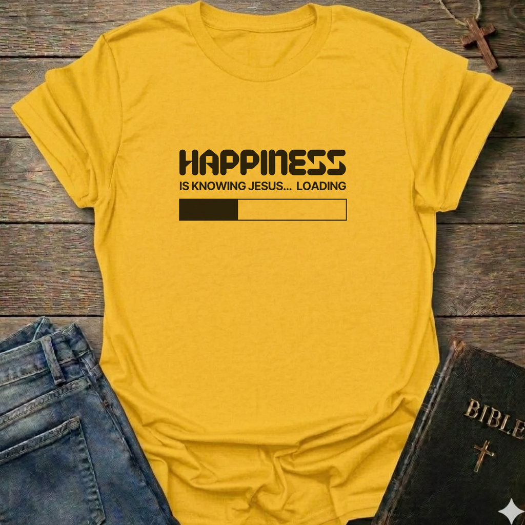 A yellow graphic t-shirt laid flat on a rustic wooden surface, styled with blue jeans and a black Bible. The t-shirt features a black printed loading bar "like a battery charging" graphic that reads 'HAPPINESS IS KNOWING JESUS.
