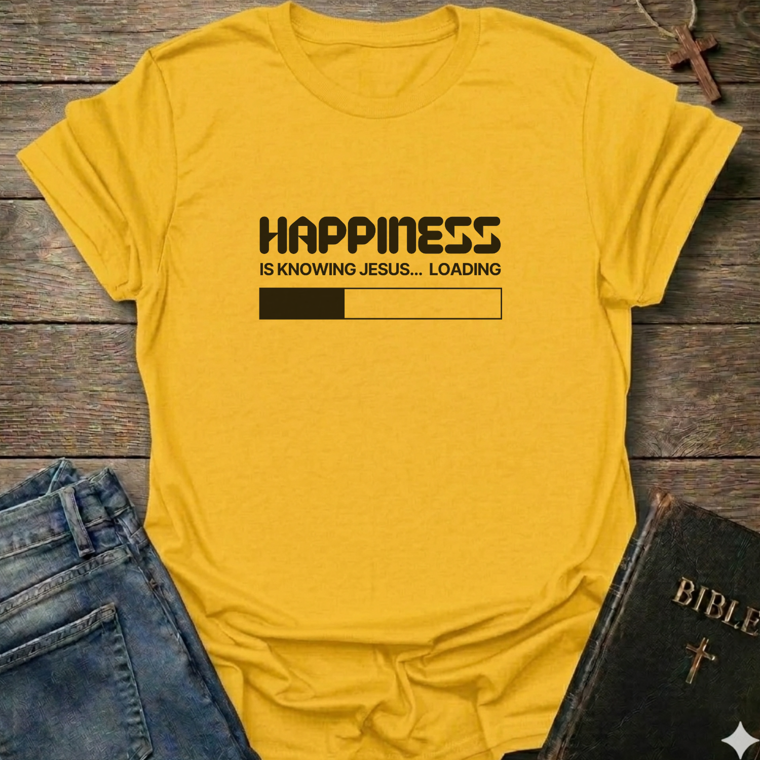 A yellow graphic t-shirt laid flat on a rustic wooden surface, styled with blue jeans and a black Bible. The t-shirt features a black printed loading bar "like a battery charging" graphic that reads 'HAPPINESS IS KNOWING JESUS.