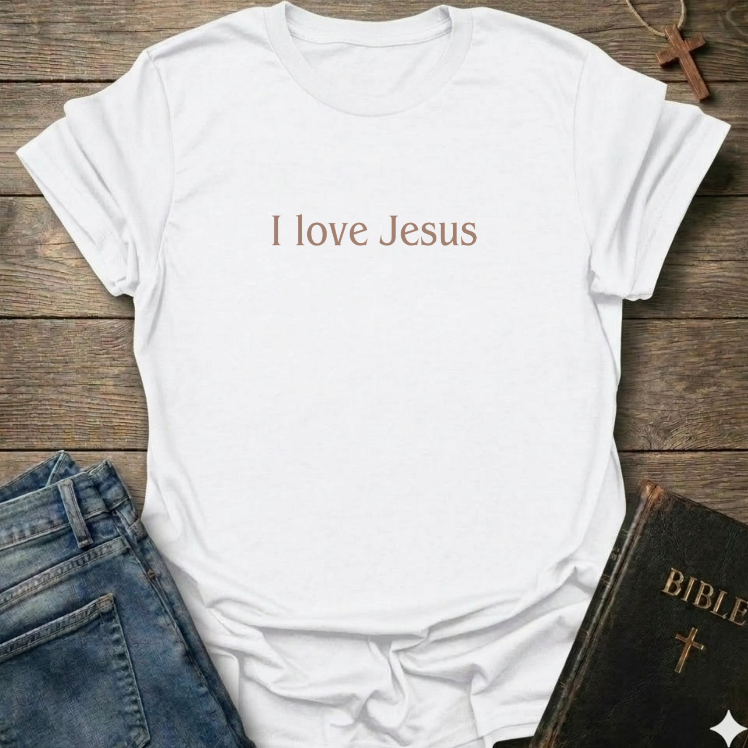 A white short-sleeve T-shirt laid flat on a rustic wooden surface, with the phrase 'I love Jesus' printed in a muted brown font across the chest. Blue jeans and a black Bible with a gold cross are partially visible as styling props.