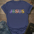 A navy blue graphic t-shirt laid flat on a rustic wooden background, styled with blue jeans and a Bible. The t-shirt features the word 'JESUS' in large, colorful letters (purple, green, orange, yellow) with the phrase 'In the Morning, Afternoon, Evening & Night' printed in white script on top of it.