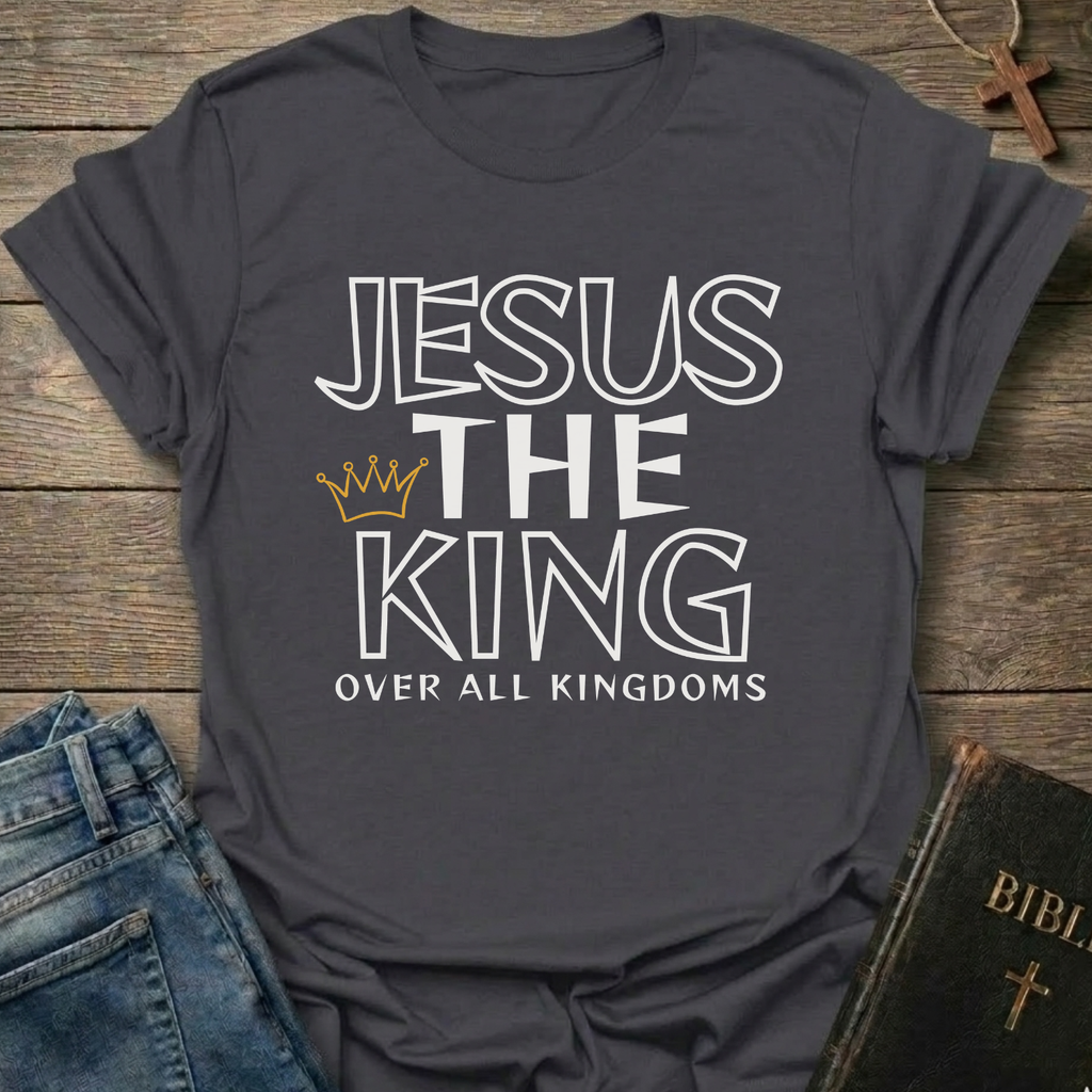 Gray t-shirt with text 'Jesus The King Over All Kingdoms'  and crown Christian bold faith apparel Faith Forward text on a wooden surface with jeans and a Bible.