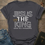 Gray t-shirt with text 'Jesus The King Over All Kingdoms'  and crown Christian bold faith apparel Faith Forward text on a wooden surface with jeans and a Bible.