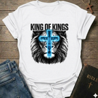White t-shirt with lion graphic and 'King of Kings' text Christian T-Shirt Revelation 19:16 bold faith apparel conversation starter on a wooden surface with jeans and a Bible.