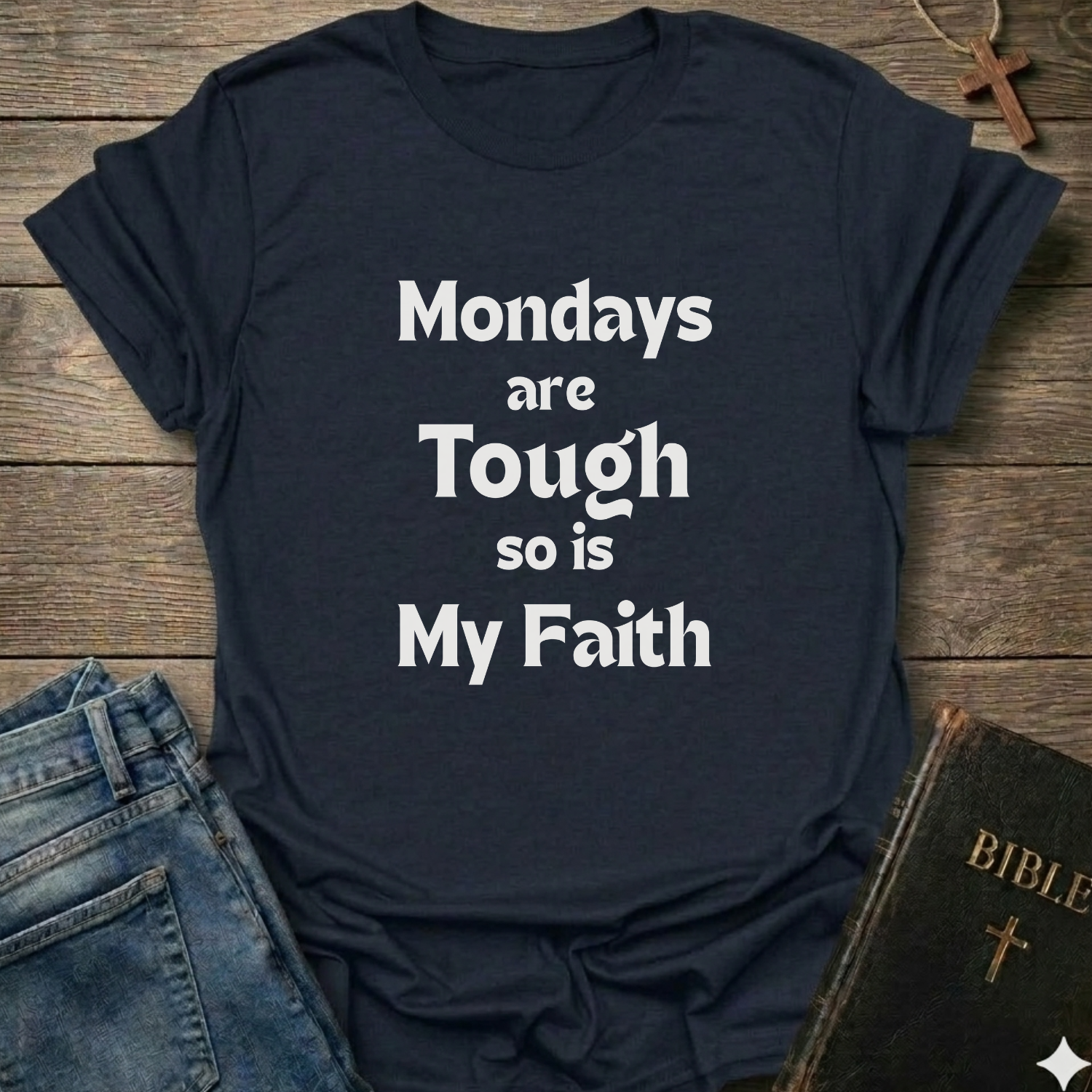 A navy blue graphic t-shirt with the text 'Mondays are Tough so is My Faith' printed in white. The t-shirt is styled in a flat-lay arrangement on a rustic wooden background, accompanied by blue jeans, a dark green Bible, and a wooden cross necklace.
