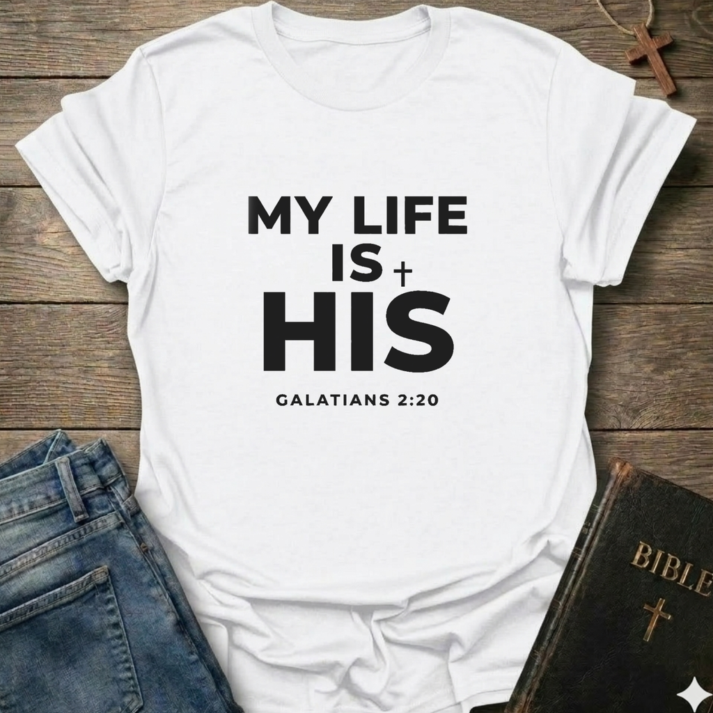 White t-shirt with 'My Life Is His'  Christian T-Shirt Galatians 2:20 bold faith apparel Faith Forward text on a wooden surface with jeans and a Bible.